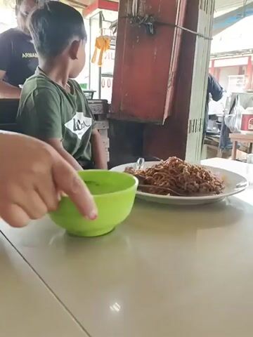 The scene shows a young boy in a green t-shirt sitting at a table in what appears to be an outdoor dining area. On the table are plates and bowls of food, including what looks like noodles and soup.  The boy is looking off to the side. In the background, there are people and structures suggesting an active street or market. The lighting is bright, indicating it's daytime.