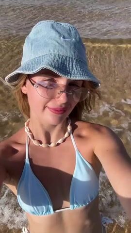 A young woman with blonde hair and a light tan is in the ocean, with waves gently lapping around her. She is wearing a light blue bikini, a light blue denim bucket hat, and sunglasses with a pinkish tint. A shell necklace adorns her neck. The water is clear and shallow, with a sandy bottom visible. The sun is shining, casting dappled light on the water and the woman. She is smiling and looking at the camera, appearing happy and relaxed. The time of day appears to be daytime, likely late morning or early afternoon given the brightness and angle of the sun. The presence of the ocean and beachwear strongly suggests a coastal or beach location. The water is calm, indicating pleasant weather conditions. The overall mood is one of leisure and enjoyment of a summer day at the beach. There is a small, subtle Nike logo on the front of her bucket hat.