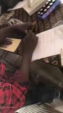 In the video, a person is drawing and coloring on a piece of white paper. They are using a pencil to draw a grid and then filling the spaces in with various colors, resulting in a rainbow-like pattern.  The person is seated, wearing a red and black checkered shirt and black jeans. The drawing surface appears to be a dark plywood surface with the word "STRONG" and "BOARD" printed repeatedly. Other objects on the surface include a container of colored pencils, a palette with paints, and a clear plastic container. There are also scattered pieces of white chalk or similar material.
