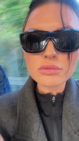 A woman wearing sunglasses and a gray coat is shown from the upper chest up, applying lip liner with a light-colored lip pencil. She is presumably on a moving train or bus based on the blurred background of trees and sunlight streaming through the window. Her nails are painted with a black and red design. She opens her mouth slightly to apply the lip liner.