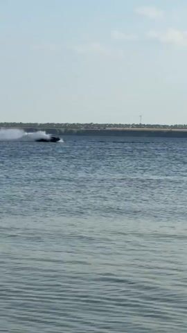 The video captures a jet ski zipping across a body of water, leaving a trail of spray in its wake. The rider, a person on the jet ski, is visible but not distinctly clear. The jet ski itself is a dark color, possibly blue or black, with a lighter colored seat.

The scene is set against a backdrop of a calm, blue sky with scattered white clouds. In the distance, a flat, arid landscape stretches out, punctuated by several wind turbines. The water appears rippled, suggesting a gentle breeze. The presence of the wind turbines and the dry vegetation on the shore indicate a rural or semi-rural setting.

The time of day appears to be daytime, likely afternoon, given the bright sunlight. The weather seems pleasant and clear, suitable for water activities. The overall mood is one of recreation and enjoyment, with the jet ski activity conveying a sense of speed and freedom.

No visible text is present in the video. The location is specified as Ukrainka, Ukraine, which aligns with the visual cues of wind turbines and the type of landscape.