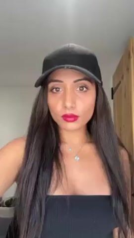 A woman with long, straight, dark hair, wearing a black strapless top, a silver necklace, a black baseball cap, and bright red lipstick is standing indoors in front of a white wall. The corner of a wooden door is visible to the right of the frame. She looks directly at the camera and runs her hand through her hair.