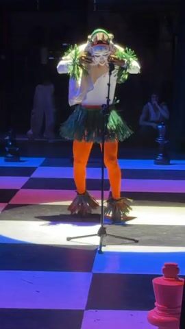 A person dressed in an unconventional costume stands on a checkered stage. The costume consists of bright orange tights, a green fringe skirt, and a large white headpiece decorated with green fringe and other colorful elements. The person is positioned behind a microphone stand. The stage is illuminated with blue and white light. Other people are visible in the background, including one seated on a chess piece, and another standing further back. Additional chess pieces are visible as well.