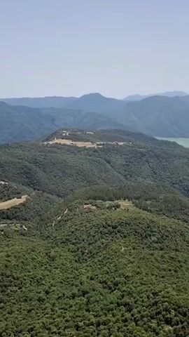 An aerial drone shot slowly pans across a vast, rolling landscape covered in dense, green forest. In the distance, several layers of hazy blue mountains recede into the horizon under a clear, pale sky. In the mid-ground, a light green body of water is visible nestled between two rounded mountain ridges. Closer to the foreground, the forest thins out to reveal a golf course with manicured fairways and winding cart paths cut into the hillsides. The golf course appears to be mostly empty. The shot is captured from a high vantage point, likely from a drone or helicopter, giving a sweeping overview of the natural and man-made elements of the environment. The bottom right corner of the frame shows a small portion of a dark brown, possibly wooden, railing or structure, indicating the viewer's perspective might be from a lookout point or a balcony. The lighting suggests it is daytime, with a bright, diffused light casting soft shadows.