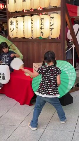Two young girls are positioned near a stall adorned with Japanese lanterns. The girl on the left, wearing a dark blue shirt and sitting, holds a green parasol and a white umbrella. The girl on the right, in a black shirt with a bow pattern and blue jeans, stands and actively fans herself with a white fan while looking towards the left. A red tablecloth covers a table on the left, holding a wooden crate. A large green, open parasol is situated behind the girl on the right. The environment appears to be an outdoor marketplace or festival, with a tiled floor and modern building architecture visible in the background.