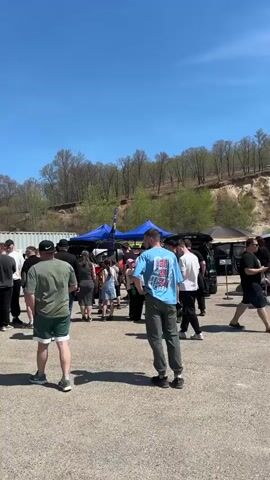 A medium shot of a parking lot filled with people and several cars. Behind them are several tents, a shipping container, and a hillside. Most of the people in the crowd are standing still and seem to be talking amongst themselves. The people in the shot seem to be of varying ages, genders, and races. The weather seems clear and sunny.