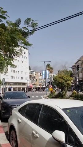 The video shows a street in what appears to be a city, with buildings on either side, palm trees, cars parked on the side of the road, and people crossing the street at a crosswalk. There are Israeli flags hanging from a lamppost. In the distance, there is what looks like smoke rising into the sky. The sky is blue with some haze. The overall impression is one of a bustling urban environment.