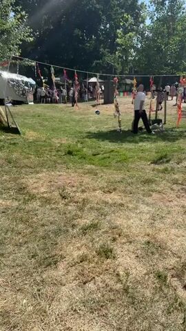 This is a first-person perspective video of an outdoor event. In the foreground, dry grass and a path are visible. As the camera moves, a man in a white shirt and dark pants is seen standing in front of a structure decorated with colorful streamers, holding a ball. Further in the background, more people are gathered, some standing and some walking, near tents and other structures. Bicycles are parked on the grass to the right. The scene appears to be a sunny day with trees and foliage in the background.
