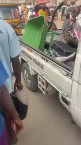 The video shows a busy street scene in what appears to be an urban area. There are people walking and standing around, some talking on cell phones. Several vehicles are visible, including a small truck with a green wheelbarrow in the back, a yellow auto rickshaw (tuk-tuk), and other cars in the distance. The truck has the words "DAVIDSON MEXUS GLOBAL RESOURCES" printed on its side. The environment seems crowded and bustling, with buildings and other structures lining the street.