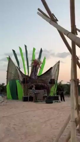 The video showcases an outdoor event featuring music and scenic views. The initial scene presents a stage constructed from natural materials like bamboo and thatch, decorated with vibrant green accents and a dragon-like head sculpture. A DJ stands behind the deck with speakers positioned around the stage. In the next shot, a bamboo structure is seen with people standing nearby. Finally, the scene transitions to a young girl standing on an elevated platform with a rope net as a railing, overlooking a landscape with flags and a stunning view of an ocean and islands. The environment suggests a peaceful, possibly tropical setting with light filtering through the sky.