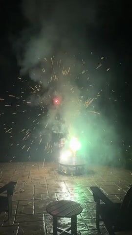 A firework is ignited on a paved patio at night. Sparks and flames shoot upwards from the firework, creating a bright explosion of light and smoke. Two chairs and a table are visible in the foreground.