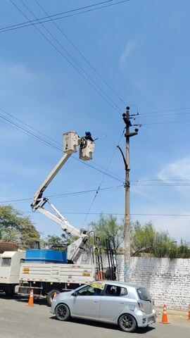 An eye-level, static shot shows a worker in an elevated platform, cleaning power lines with a water hose. The worker is stationed in a white bucket lift attached to a white truck with a blue container in the back. The lift extends towards the top of a power pole located on the right side of the frame. The worker, clad in dark clothing, sprays a stream of water directly at the insulators on the power pole, presumably to clean them. The sky is clear blue with scattered clouds. A silver car is parked on the left side of the frame. Orange traffic cones are placed around the base of the truck. Another worker, wearing an orange safety vest and hard hat, stands on the ground near the truck and power pole. In the background, there is a white brick wall and some greenery.