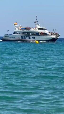 This is a video of a boat named "NEPTUNE" sailing on a clear blue sea under a bright sunny sky. The boat appears to be a passenger vessel, possibly a tour boat, as there are many people visible on its upper deck, some standing and others seated. The text "DÖFI JET BOATS" and "VISIO SUBMARINA" are visible on the side of the boat, suggesting it offers submarine tours. A Spanish flag is flying from the stern of the boat. The water is calm with gentle ripples, indicating pleasant weather. Several yellow buoys are scattered in the sea, marking a path or an area. In the foreground, a person is swimming in the water, looking towards the boat. The scene is set in a coastal area, likely a beach resort given the presence of swimmers and tour boats. The time of day appears to be midday, judging by the strong sunlight and clear sky. The overall mood is one of leisure and enjoyment of a summer day at sea. The boat's registration number "PM-1-138" is visible.