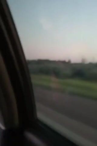 The video shows a blurred view from inside a vehicle, looking out a window. The outside scenery consists of a road, green fields, and trees. The sky appears to be a pale blue with hints of pink, suggesting either dawn or dusk. The interior of the car is dark and out of focus, with the edge of the window frame visible on the left side. There are no people or specific objects clearly identifiable outside the vehicle due to the blurriness. The overall impression is of movement, as if the footage was captured while the vehicle was in motion.