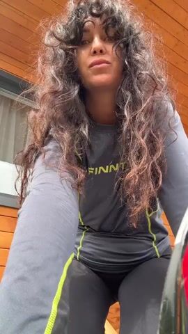 A woman with long, curly dark hair, wearing a grey long-sleeved shirt with neon green accents and black leggings, is in a low crouch position. She is outdoors, in front of a wooden wall. She moves her hands, running them through her hair and then gesturing outward. Her facial expression changes from neutral to a slight smile and then a playful pout.