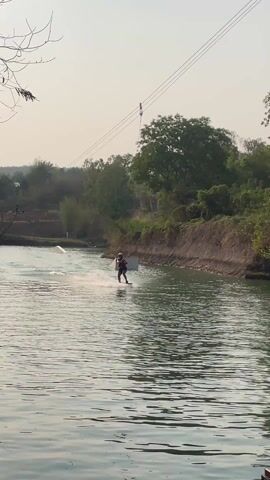 The video shows a wakeboarder being pulled along a river by a cable system. The person is wearing a helmet and life vest and is being pulled from right to left across the frame. The river is surrounded by trees and vegetation. There are two rectangular ramps on the left side of the river. The sky is hazy.