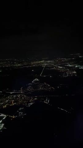 The video shows an aerial view of a city at night. City lights shine through the darkness. There are some clouds at the top of the frame.