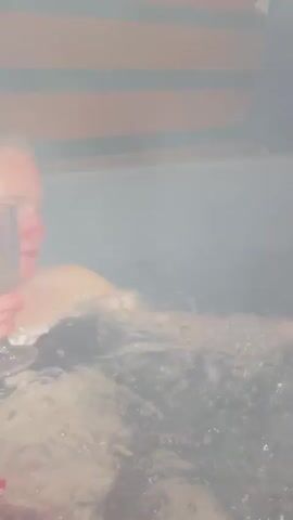 The video begins with a close-up shot of water bubbling in a dark hot tub, illuminated by blue light. A person's arm is partially visible, submerged in the water. The view then shifts to show the person getting out of the hot tub and standing near the edge. In the background, there is a dark, misty outdoor scene. The scene is framed by a wooden structure with string lights overhead, and a dark road is visible in the foreground. Distant lights and shadows suggest a vehicle and some structures in the hazy background.