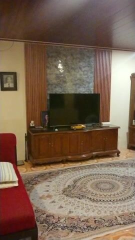 The video captures a cozy living room featuring a blend of traditional and contemporary elements. A red tufted sofa sits on a patterned rug, facing a wooden TV stand with a flat-screen TV. The wall behind the TV is adorned with decorative panels. A wooden display cabinet stands nearby. The room is lit by overhead lights, highlighting the wooden ceiling and window frames. The overall ambiance suggests a comfortable and inviting space.