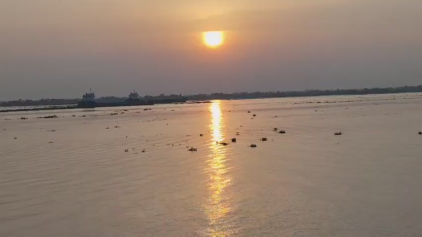 The scene is a body of water, likely a river or wide stream, at sunset. The sun is low in the sky, partially obscured by a hazy atmosphere, casting a bright golden reflection on the water's surface. Several clumps of vegetation float on the water.  In the distance, a silhouette of land with trees is visible, along with two large ships or barges anchored near the shore. Ripples spread across the water, and the sky is a pale, muted color.
