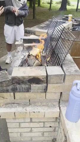The video shows a man standing near a brick barbecue pit with a fire burning inside. The man is wearing a grey sweater, white shorts, and white sneakers. He is holding a white cup and appears to be looking at his phone. A grill grate is positioned over the fire. To the right of the barbecue pit, a large blue water bottle is visible. In the background, trees and other barbecue pits can be seen. The scene appears to be outdoors, possibly in a park or campground.