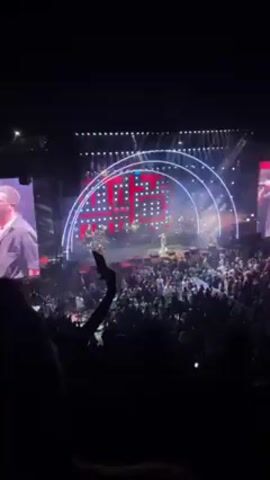 The video shows a concert with a band on stage, including a lead singer, drummer, and guitarist. The stage has a large LED screen behind the band, displaying various colorful patterns and designs. There are stage lights. A large crowd of people is in front of the stage. Many are holding up their phones to record the concert. Some people are raising their hands in the air and dancing. The video is taken from the perspective of someone in the crowd.