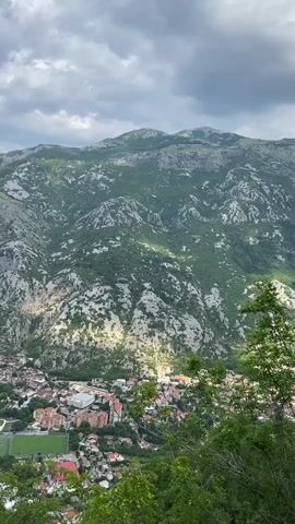 This is a landscape video looking down on a town nestled at the base of a steep, rocky mountain. The mountain is covered in sparse green vegetation and is partially illuminated by sunlight breaking through dramatic, cloudy skies. Below, the town features red-tiled roofs, a large green sports field, and a body of water with boats. The scene appears to be a scenic overlook, with lush green foliage in the foreground suggesting the viewpoint is from a higher elevation. The time of day seems to be late afternoon or early evening, given the lighting and cloud cover. The location is identifiable as Škaljari, Montenegro, based on the distinct topography and the presence of a prominent bay or fjord, characteristic of the Bay of Kotor. There are no people or discernible objects that suggest a specific event. The overall mood is one of natural beauty and awe, with the grandeur of the mountains dominating the scene.