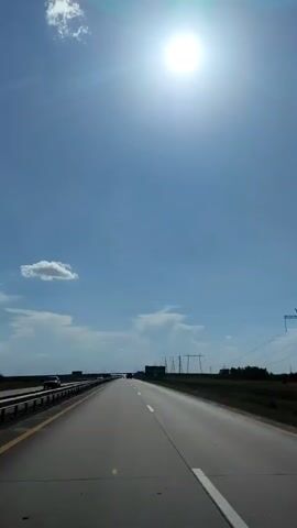The video shows a first-person perspective of driving on a highway on a sunny day. The sun is bright in the upper center of the frame, and there are a few scattered clouds in the blue sky. The road ahead is empty, with lane markings visible. To the left of the road, there is a barrier and some vegetation. To the right, there are trees and power lines. A few cars are visible in the distance on the highway. The overall impression is of a clear, bright day with unobstructed visibility.