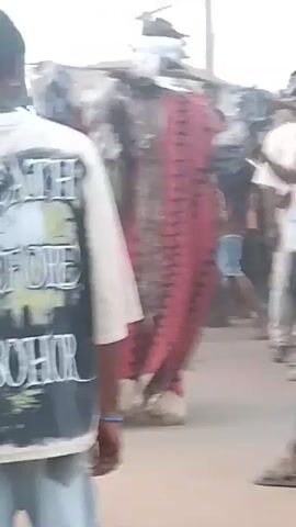 The video shows a group of people gathered on a street. In the center is a figure wearing a tall, elaborate costume with red fabric and black zig-zag patterns. The figure is surrounded by other people, some of whom are taking photos or videos with their phones. The crowd includes people wearing traditional clothing, such as white robes and caps. The setting appears to be a public street, with utility poles visible in the background. The overall atmosphere seems festive or ceremonial.