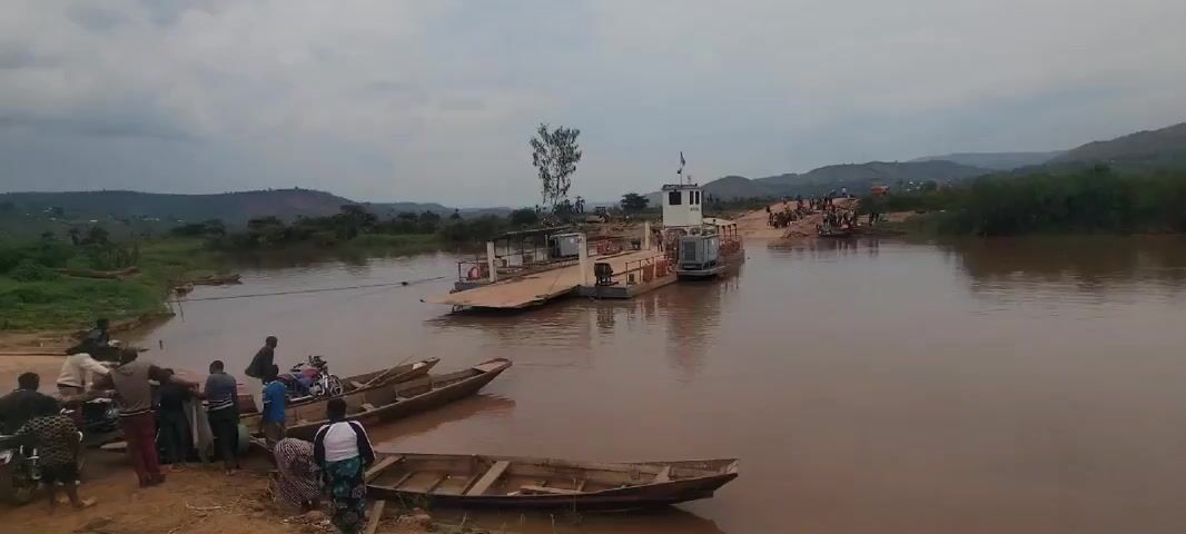 The video shows a river crossing via a small ferry. Several people are standing near the river bank next to wooden boats. One person is on a motorcycle. The ferry is mid-river, attached to a cable spanning the river. The ferry has a small structure on it, possibly a control area or small cabin. Other people are seen on the opposite bank, waiting to board or having just disembarked. The water is brown, likely due to sediment. Hills can be seen in the background, and the sky is overcast.