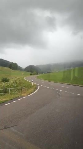The media file shows a view from a moving vehicle, likely a bus or car, traveling on a winding road through a rural landscape. The road is paved with asphalt and has white dashed lines marking the lanes. On the left side of the road, there are green fields with wildflowers and a wooden fence. Further back, hills covered with trees rise up, and some patches of fog or clouds obscure the tops of the trees. A yellow diamond-shaped road sign with a black symbol is visible on the left side of the road.

The weather appears to be overcast and possibly rainy, with a heavy layer of gray clouds in the sky. The overall mood is calm and serene, with the natural beauty of the landscape being the main focus. There are no people or animals visible in the frame. The time of day is not explicitly indicated but the dim light suggests it might be morning or late afternoon. The visual style suggests the footage was captured on a mobile device, possibly through a window, as some reflections are visible on the surface. The location is identified as Petit-Val, Switzerland.