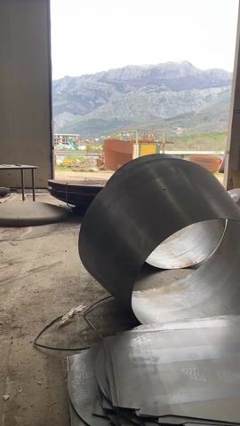 From inside a large industrial building, a view looks out through a wide opening onto a construction yard. Several large pieces of rounded metal sit on the concrete floor in the foreground. Just outside the opening, there is a stack of large bowl-shaped metal structures. Outside in the yard, other metal structures are visible, along with a small yellow building and construction vehicles. In the distance, a range of large, rocky mountains forms the backdrop. The sky is overcast.