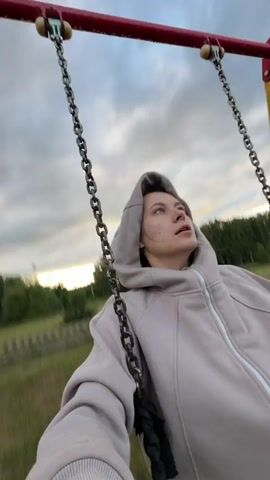 A low-angle, first-person perspective video shows a person wearing a light grey hooded sweatshirt and pants sitting on a swing. The swing has metal chains and a black rope seat. The background consists of a cloudy sky and a tree line in the distance. The person is looking upwards and to the side. The swing appears to be on a playground with a red and yellow frame. The video captures the motion of swinging back and forth.