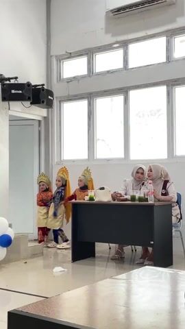The video shows a group of four young children in traditional Indonesian attire, likely for a performance, standing on a raised platform to the left. They are wearing ornate headdresses and colorful costumes. To the right, behind a black table, sit two women wearing hijabs. They appear to be judges or audience members, with drinks and a bag on the table in front of them. The setting seems to be an indoor venue, possibly a school or community hall, with large windows behind the table and an air conditioning unit visible on the wall. The lighting suggests daytime. The children appear engaged in their performance, while the women seem attentive and possibly discussing the performance. Balloons, one blue and two white, are visible to the left of the children. The overall mood appears to be one of cultural celebration and performance.