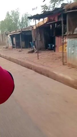 The video shows a person in a red and black shirt riding in a vehicle down a street. The camera is positioned to the side of the vehicle, capturing the view of the roadside. The street is paved and runs alongside a row of small, makeshift structures that appear to be shops or stalls, some are open and others are closed. The structures are constructed from corrugated metal and wood. Behind the structures is a tall, concrete wall that is partially broken in places. Trees can be seen above the wall. The sky is overcast.