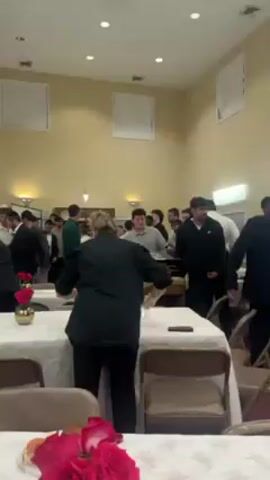 The video depicts a room with people setting up tables for an event. The room has a beige and white color scheme with rectangular white panels on the ceiling. A group of people, mostly men, are gathered around the tables, moving and talking. Some are wearing traditional Jewish attire, including yarmulkes and suits. A woman in a black shirt is arranging items on a table in the foreground, near a floral centerpiece with red roses. There's a gilded box-like structure on one of the tables. The tables are covered in white tablecloths, and brown folding chairs are placed around them. The scene suggests preparations for a gathering, possibly a religious or community event.