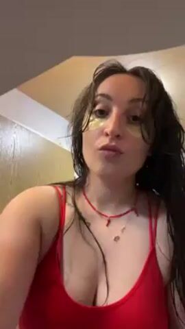 Here is a description of the video.
The video shows a woman in a red top, with eye patches on, wet hair and a red beaded necklace. She is looking at the camera and pouting slightly.  She then tilts her head to the left and then back.  The background is of a room with wood panelling.