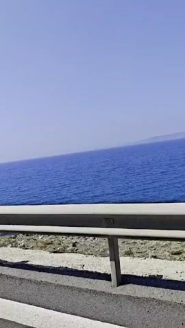 The video shows a view of the sea from a roadside with a guardrail. The sea is a deep blue color and stretches to the horizon where the sky is a lighter shade of blue. A road sign indicating a curve is visible in the left foreground. The road appears to be asphalt with white lines marking the edge. The guardrail is silver. The landscape between the road and the sea is sparse with low-lying vegetation.