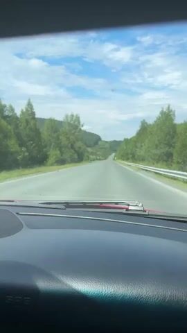 The video shows a first-person perspective of driving on a highway. The road stretches ahead, flanked by lush green trees and rolling hills under a bright blue sky with scattered clouds. A white guardrail is visible along the right side of the road. The dashboard of the car is in the foreground, with windshield wipers resting on the windshield. The scene appears to be a sunny day, possibly in a rural or mountainous region. There are no other vehicles or people visible in the frame.