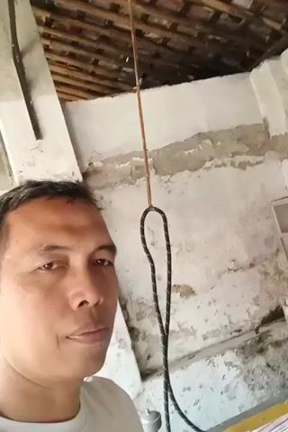 A man in a white t-shirt is holding an iron in his right hand, posing for a selfie. He is indoors in a room with a simple, rustic appearance. The walls are rough and show signs of wear and tear, with peeling plaster and some discoloration. Above, the ceiling appears to be made of wooden beams. To the right of the man, there is a white cabinet filled with various kitchen utensils and containers. A wire hangs from the ceiling, possibly part of a lamp or fan. The man's expression is neutral, and he is looking directly at the camera. The overall impression is one of a domestic setting, possibly a kitchen or storage area.
