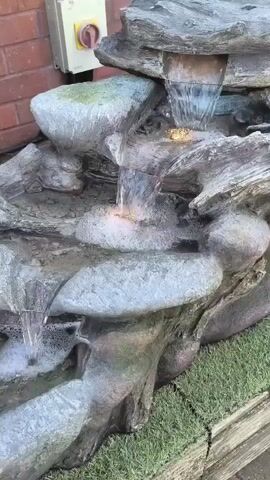 A close-up, eye-level, medium shot video shows a decorative water fountain made to look like rocks and driftwood. Water cascades down several levels of the fountain, creating small waterfalls and foam at the bottom. The fountain is situated outdoors, with a brick wall in the background and artificial grass on the ground. A white electrical box with a red switch is visible on the brick wall. The lighting is natural, suggesting it is daytime. The camera pans slightly, showing the detail of the faux wood and stone textures of the fountain.