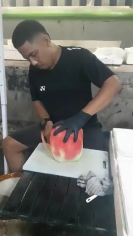 A man wearing a black t-shirt and black gloves is sitting at a table and cutting a watermelon with a knife. He is holding the watermelon with his left hand and cutting it with his right hand. The watermelon is red and white and is on a white cutting board. There is a checkered cloth on the table next to the cutting board. The background appears to be a market or food stall with shelving and boxes.