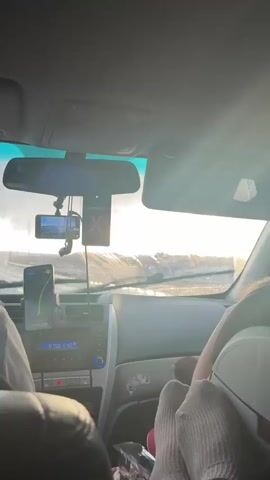 From inside a car, the video shows a view through the windshield towards the horizon. The weather appears wet, with raindrops visible on the glass. A black dashcam is mounted near the rearview mirror, along with a black air freshener. A smartphone in a holder to the left displays a navigation app. The car's radio shows 87.50 FM. Sunlight filters through the clouds on the horizon. The landscape includes industrial structures. A person wearing a hooded sweatshirt sits in the driver's seat.