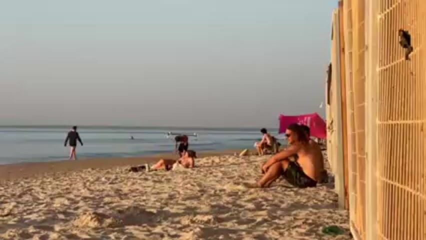 The scene is a sunny beach with several people scattered across the sand. At the shoreline, one person is walking into the water, while another is holding a long, thin object. Closer to the foreground, two people are lying on the beach, sunbathing. Further along the beach, a few people are sitting near a pink tent or windbreaker. A shirtless man with long hair sits in the foreground, leaning against a structure made of vertical slats. The overall lighting suggests it's either early morning or late afternoon. The sky is a pale, uniform color, indicating a somewhat hazy atmosphere.