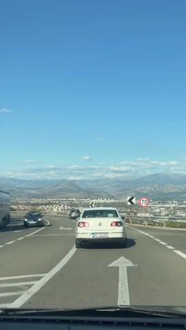 The video shows a view from inside a vehicle driving on a highway. The road curves to the left, and there are lane markings and arrows on the road. A white Volkswagen Passat is in front of the vehicle, and other cars are visible in the distance. On the right side of the road, there are guardrails, a speed limit sign indicating 70, and a chevron sign pointing to the left. In the background, there is a city spread out below, with mountains and a clear blue sky with some clouds. The sun is shining, suggesting it is daytime.