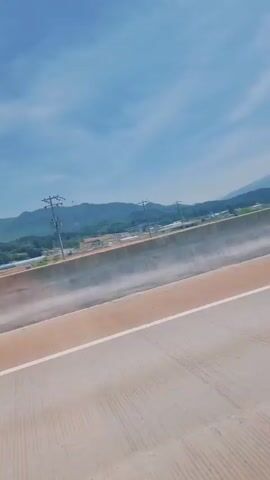 The video shows a view from a car driving on a highway. The sky is blue with some clouds. In the distance, there are green mountains. A concrete barrier and a metal guardrail are visible alongside the road. Trees and power lines dot the landscape on the other side of the guardrail.