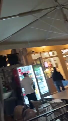 The video shows an outdoor nighttime scene. First, it focuses on a man in a white shirt standing in front of a lit refrigerator inside a building. The view is partially obstructed by what appears to be a table in the foreground. Then, the camera pans to show parked motorbikes, one with a red delivery box attached. Finally, the camera moves quickly, blurring the lights and outlines of buildings and trees as if moving through the street at night.