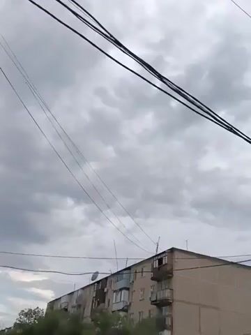 The video shows a cloudy sky with electrical wires in the foreground. A building, possibly an apartment complex, is visible below the wires. The sky is overcast with gray clouds, and there is no visible sunlight. The electrical wires are strung across the frame, creating a sense of depth and perspective. The building appears to be residential, with balconies and windows. The overall scene is mundane and lacks any specific activity or action.