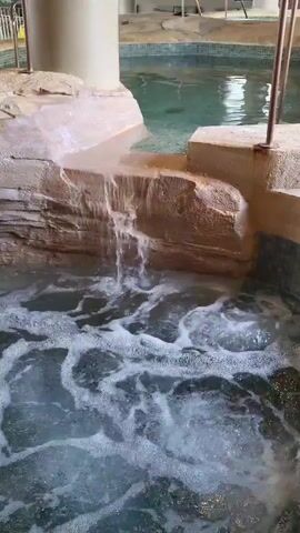The video shows a hot tub or jacuzzi with bubbles forming on the surface of the water, with a small waterfall feature flowing from a faux rock formation into the tub. The faux rock formation is light brown, and the water in both the tub and the higher pool that feeds the waterfall appears clear and slightly green. Beyond the tub is a tiled pool area and a building interior with a railing, windows, and indoor plants. A person walks by in the background behind the railing. The overall setting suggests a resort or spa.