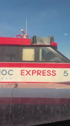 The video captures a view from inside a moving vehicle, looking out at a red and white boat labeled "EXPRESS 5" passing by. The boat has windows and is docked or moving slowly in a harbor. Beyond the boat, other vessels are visible, along with a coastline and buildings in the distance under a clear blue sky. The scene appears to be a sunny day.